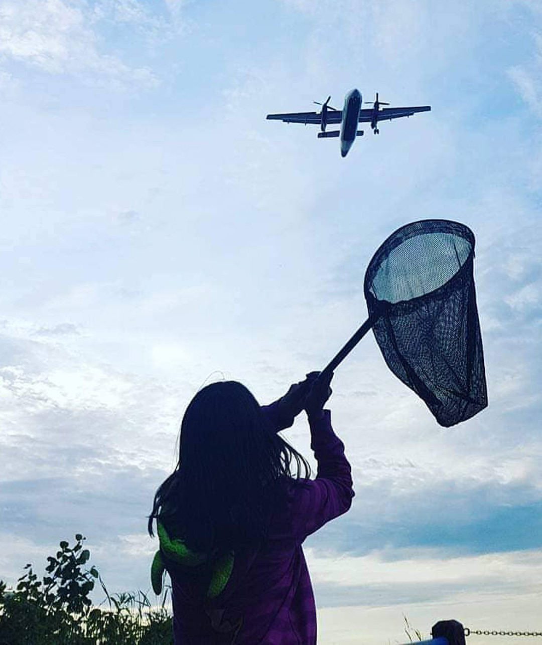 Little girl reaching her butterfly net up to an airplane in the sky