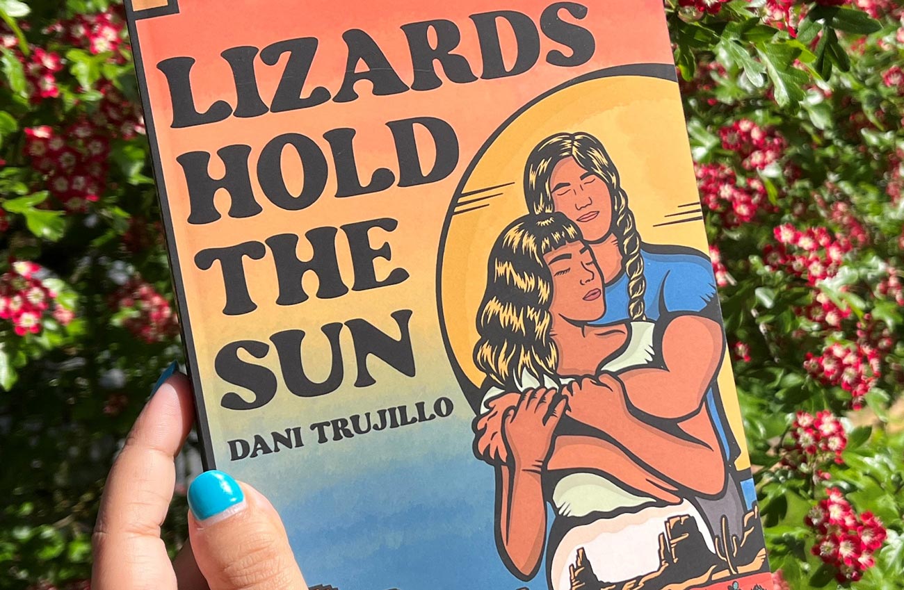 close view of the cover of Lizards Hold the Sun held in hand against a background of flowering shrubs