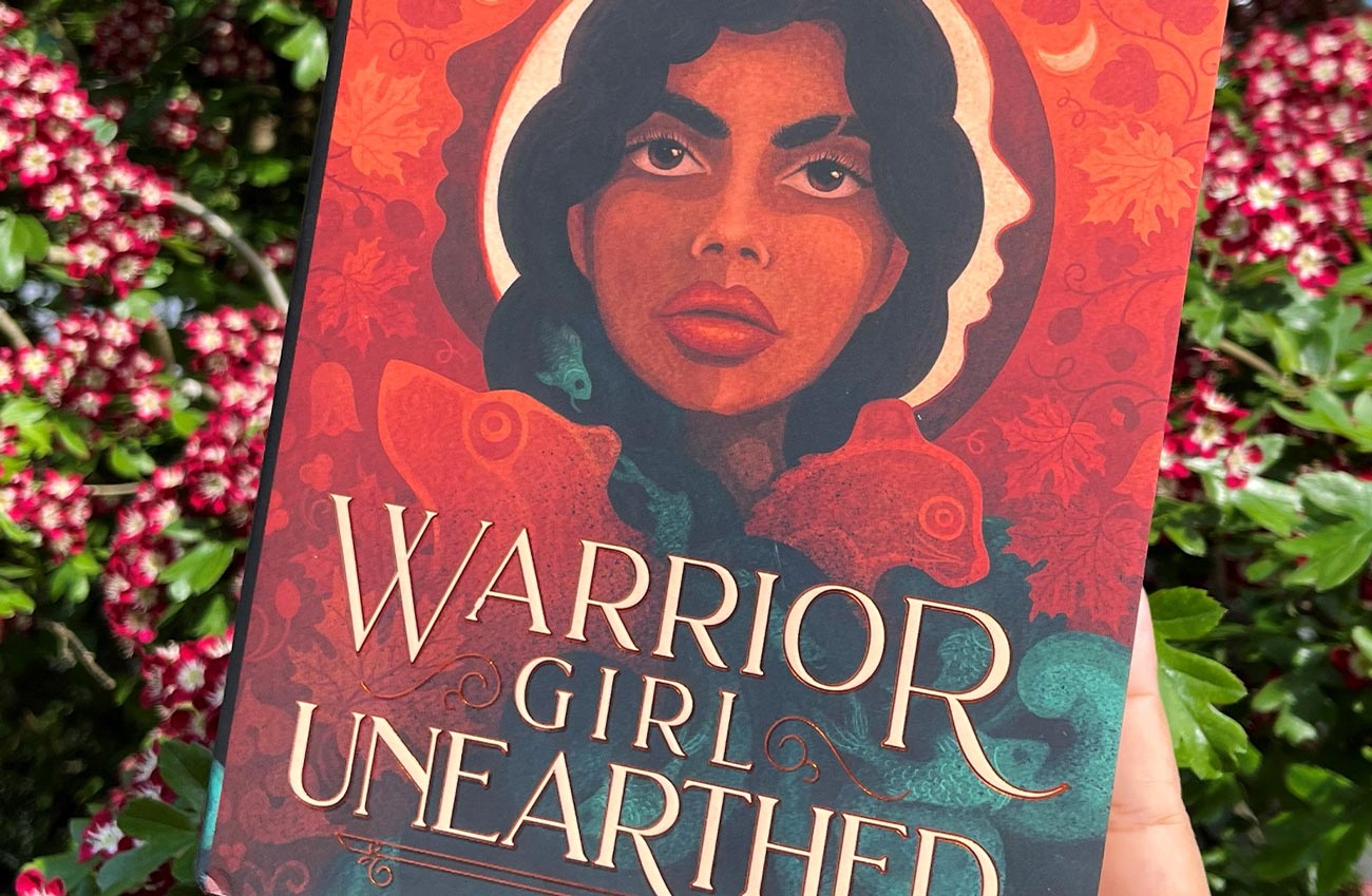 close view of the cover of Warrior Girl Unearthed held against a background of flowering shrubs