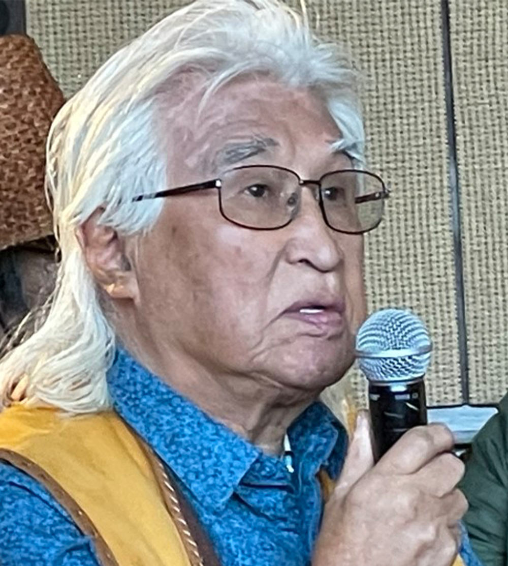 an older man with a white mullet, wearing glasses, a bright blue shirt and a tan vest looks off camera while speaking into a microphone