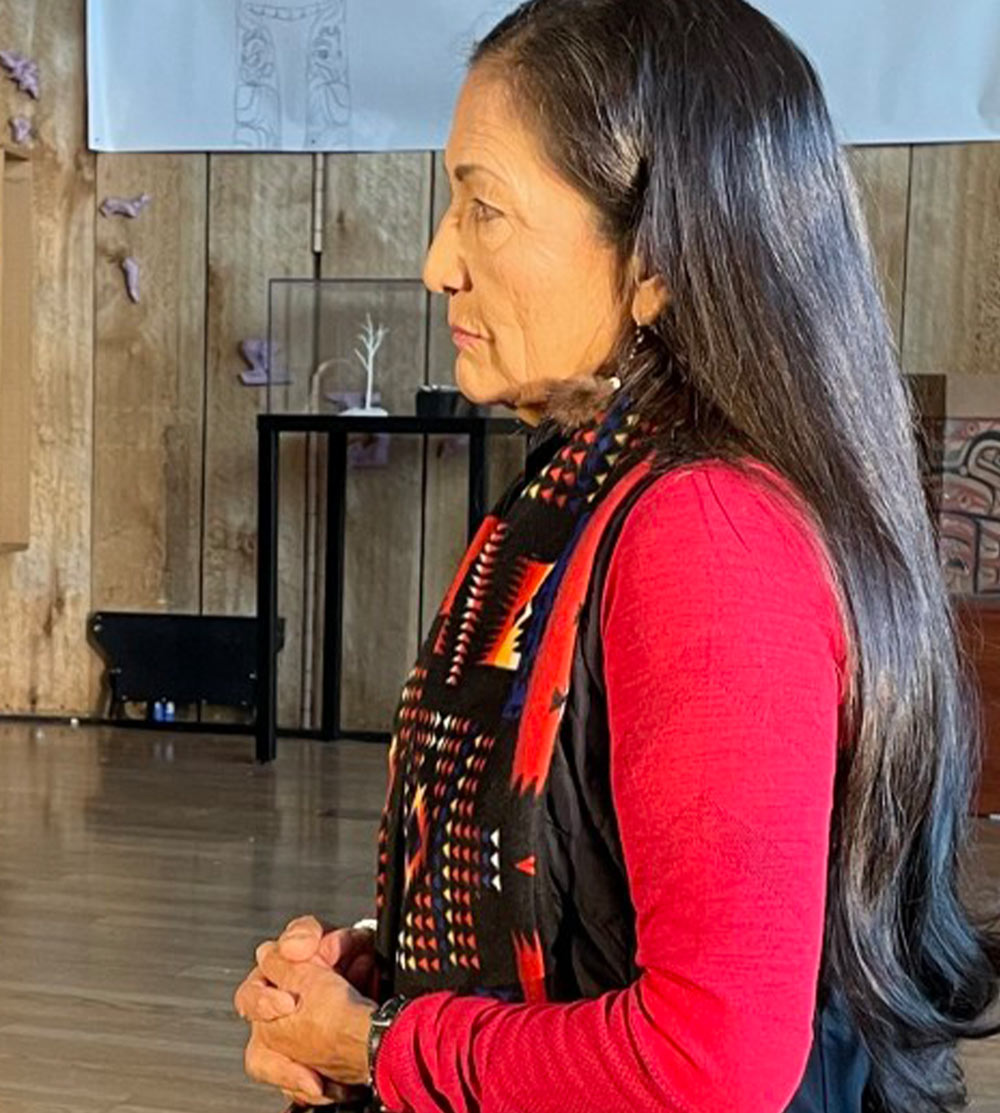 profile view of a woman whit long black hair and waring a patterend scarf and a red long sleeve shirt, she looks off camera with a look of listening