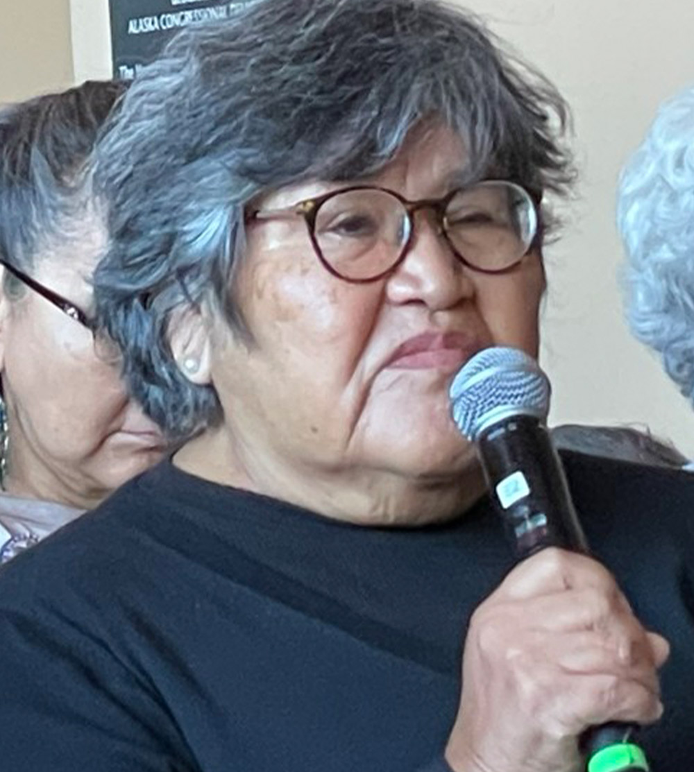 an older woman with short salt and pepper and wearing glasses and a black shirt holds a microphone to her face