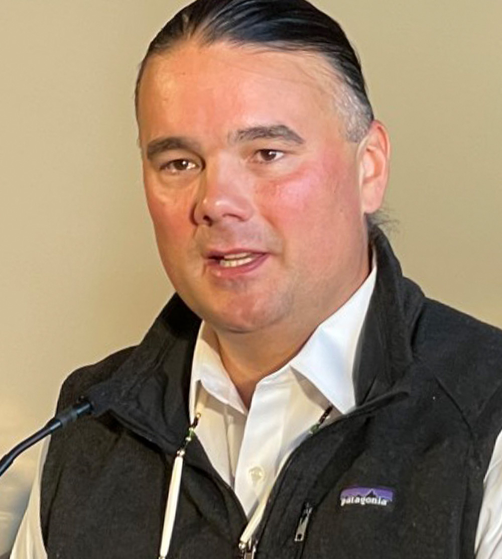 a man with his hair pulled into a ponytail wears a white button up shirt, a vest and a serious expression as he speaks into a podium microphone