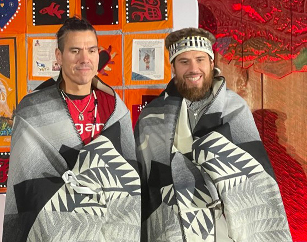 two men bearing facial resemblance, one bearded and wearing a headdress, wear traditional Haida blankets and smile for a photo