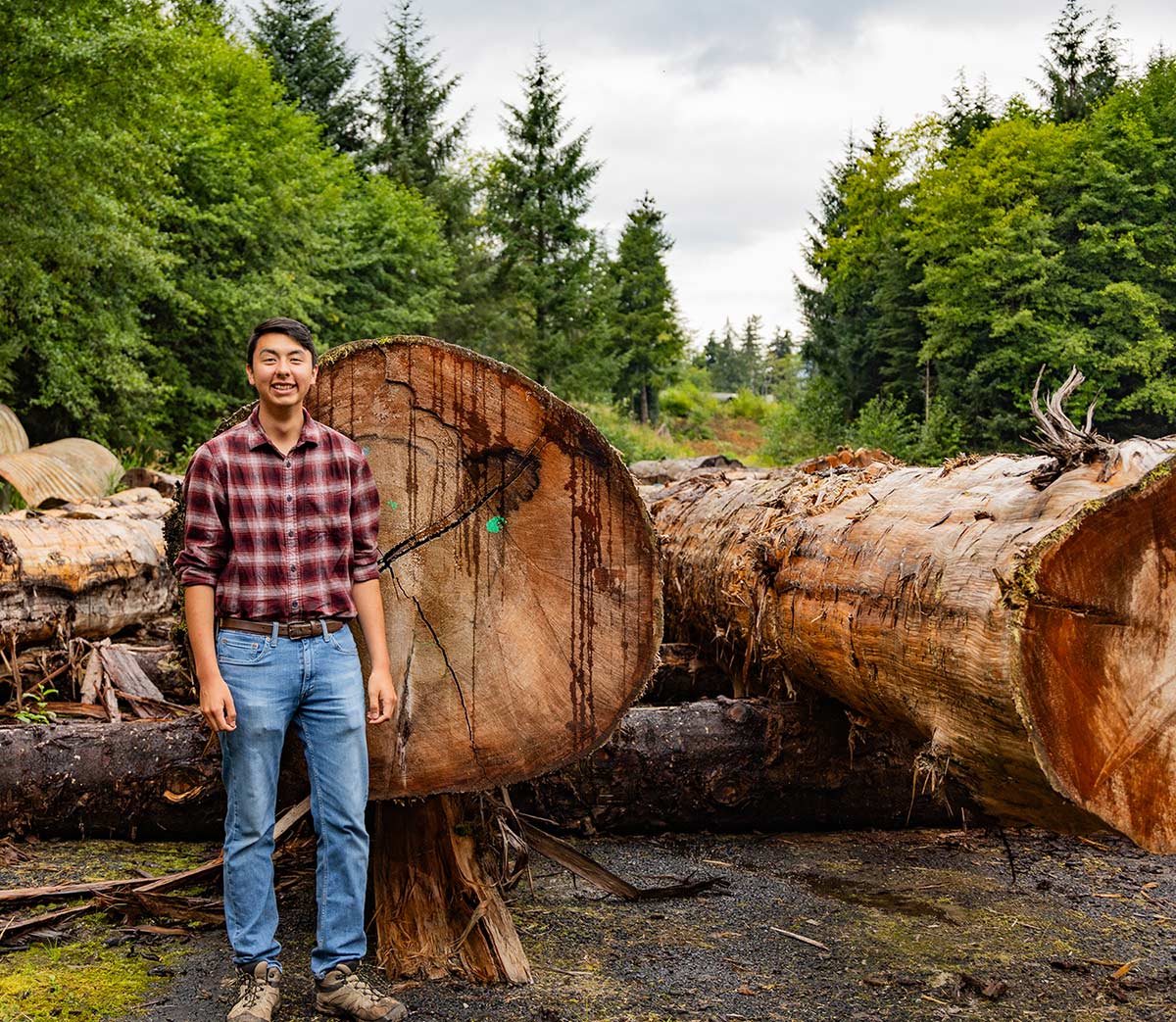 Samuel Hiratsuka in a red flannel and denim jeans standing in front of  harvested trees